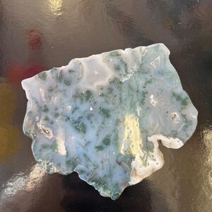 Puede incluir: Una rebanada de &aacute;gata verde y blanca con un patr&oacute;n &uacute;nico de remolinos y l&iacute;neas. La &aacute;gata es una piedra natural y tiene una superficie lisa y pulida.