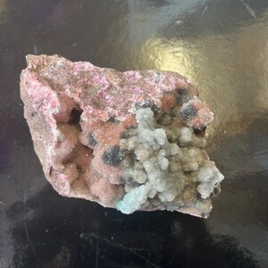 May include: A pink and white crystal cluster with a rough, brown exterior. The crystal cluster has a variety of textures and shapes, including smooth, rounded crystals and rough, jagged crystals.