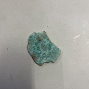 Puede incluir: Una piedra pulida de forma irregular, de color azul claro y verde, sobre un fondo blanco.
