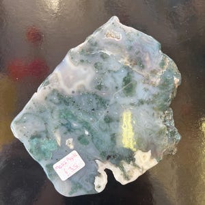 Puede incluir: Una losa de &aacute;gata verde y blanca con una forma natural irregular. La losa tiene una superficie lisa y pulida y presenta una variedad de patrones verdes y blancos. La etiqueta de precio dice "Moss Agate &pound;3.5".
