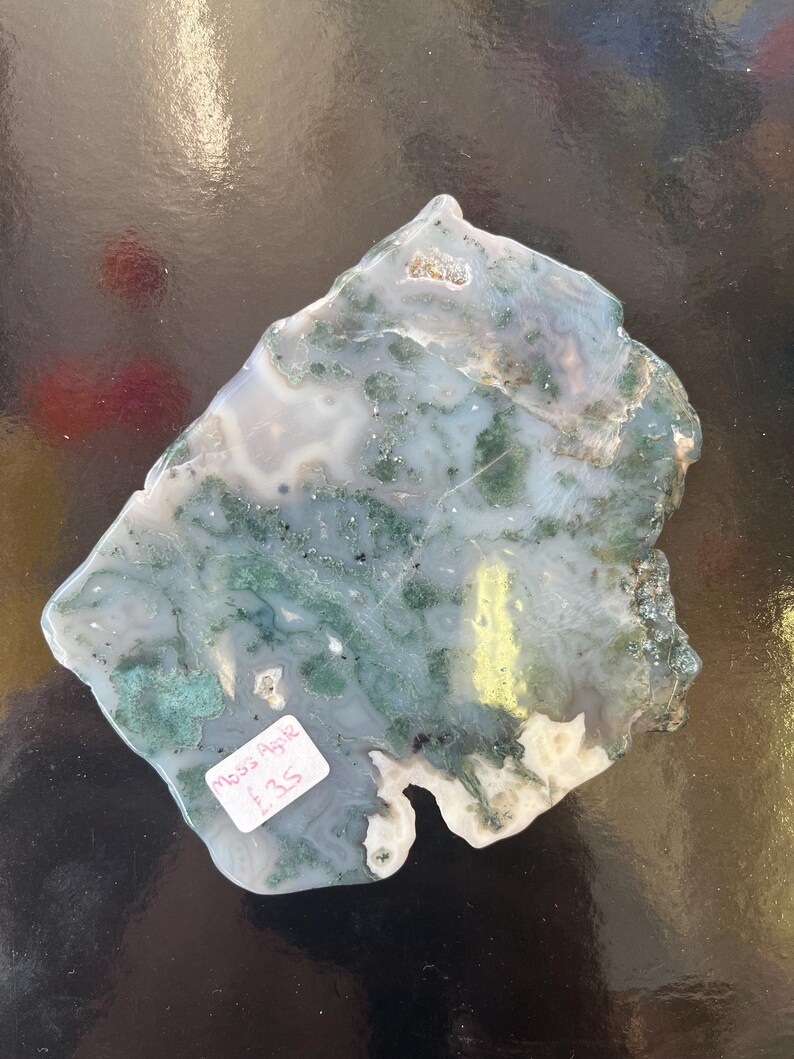 Puede incluir: Una losa de &aacute;gata verde y blanca con una forma natural irregular. La losa tiene una superficie lisa y pulida y presenta una variedad de patrones verdes y blancos. La etiqueta de precio dice "Moss Agate &pound;3.5".