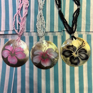 May include: Three necklaces with shell pendants featuring painted hibiscus flowers. The necklaces are made of pink, white, and black beads.
