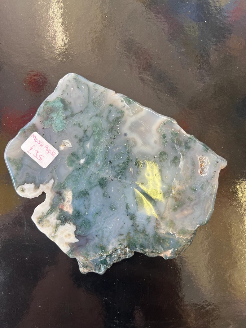 Puede incluir: Una losa de &aacute;gata verde, blanca y marr&oacute;n con una forma natural irregular. La losa mide aproximadamente 25 cm de largo y 18 cm de ancho. La &aacute;gata tiene una variedad de patrones y colores, incluyendo verde, blanco, marr&oacute;n y negro. La losa est&aacute; sobre una superficie negra. Moss Agate &pound;35