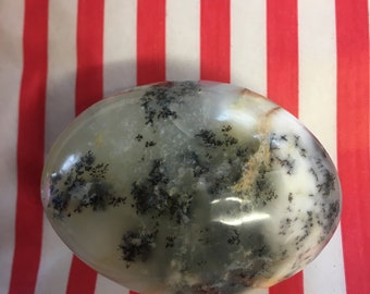 May include: A smooth, white and gray agate stone with black dendritic patterns. The stone is oval shaped and has a polished finish.