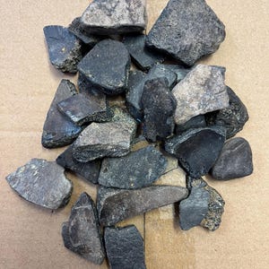 Könnte beinhalten: Eine Ansammlung unregelmäßig geformter, dunkelgrauer und schwarzer Steine. Die Steine haben raue, strukturierte Oberflächen und sind unterschiedlich groß. Einige Steine haben hellere graue Flecken.
