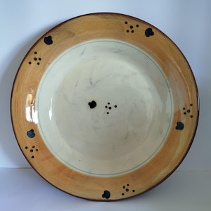 Può includere: Un grande piatto rotondo in ceramica con un bordo largo di colore marrone chiaro e un centro bianco. Il piatto è decorato con piccoli punti neri e alcune forme nere più grandi intorno al bordo.