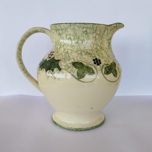 Könnte beinhalten: Eine weiße Keramikkanne mit grünem und braunem Blumendesign. Die Kanne hat einen breiten Boden und einen Henkel.