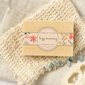 May include: A bar of handmade soap with a floral design and the text "lazy morning" on a white label. The soap is on a white mesh bag and a white surface.