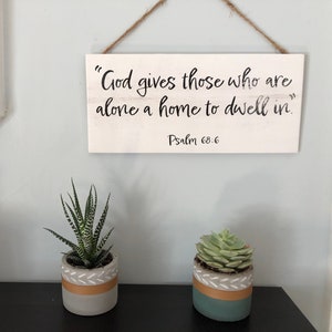 Op de afbeelding: Een wit houten bord met de tekst "God geeft degenen die alleen zijn een huis om in te wonen. Psalm 68:6" hangt aan een lichtblauwe muur. Drie kleine potplanten met vetplanten staan op een zwart plankje onder het bord.