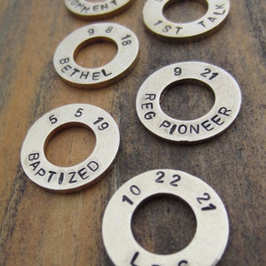 May include: Six brass rings with engraved text and numbers. The rings are on a wooden surface. The text on the rings includes "Baptized", "Bethel", "1st Talk", "Reg Pioneer", "LDC", and "Comment 2012".