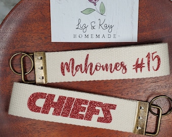 May include: Two white canvas keychains with red glitter lettering. The top keychain says "Mahomes #15" and the bottom keychain says "CHIEFS".