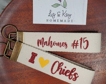 May include: Two white canvas keychains with red glitter lettering. One keychain says "Mahomes #15" and the other says "I love Chiefs". The keychains are on a wooden surface with a business card that says "Liz & Kay Homemade".