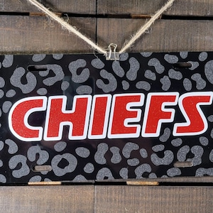 May include: A black license plate with a gray leopard print pattern and the word "CHIEFS" in red glitter letters with a white outline. The plate is hanging from a piece of twine.
