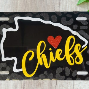 May include: Black license plate with a glitter leopard print pattern. The center features a white outline of a state, a red glitter heart, and the word "Chiefs" in yellow script.