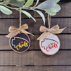 May include: Two round wood slice ornaments with jute twine hangers and burlap bows. One ornament is black with a white arrowhead outline, a red heart, and the yellow text Chiefs. The other is white with a black arrowhead outline, a gold heart, and red text Chiefs.