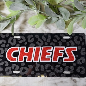May include: A black license plate with a leopard print pattern and the word "CHIEFS" in large red glitter letters with a white outline. The plate has four mounting holes.