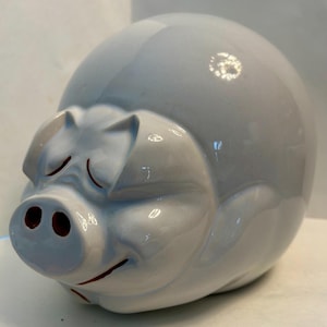 May include: A glossy, light blue ceramic piggy bank shaped like a stylized pig. The figure features a smooth, rounded body, pointed ears, and a snout with two dark red nostrils. A thin, dark red line forms a subtle, closed-eye expression on its face.