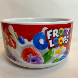 May include: A white ceramic bowl with a red exterior featuring the text "Froot Loops" and colorful cereal pieces. The bowl has a white interior and is filled with a splash of milk and various colored cereal pieces.