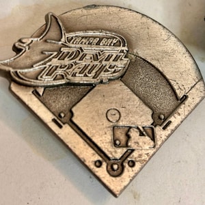 May include: A silver-toned metal baseball-themed pin featuring the Tampa Bay Devil Rays logo. The pin is shaped like a baseball diamond with the MLB logo and team name prominently displayed.