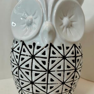 May include: A white ceramic owl figurine with a geometric black pattern on the lower half. The owl has large, round eyes and a pointed beak. The design is modern and decorative.