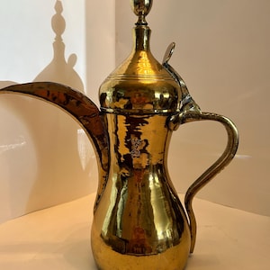 May include: A traditional brass coffee pot with a rounded body, a long spout, and a hinged lid with a decorative finial. The pot has a hammered finish and a polished shine.
