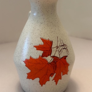 May include: A speckled, off-white ceramic pitcher with a handle. The pitcher features a design of three orange maple leaves and a sprig of seeds. The pitcher has a narrow neck and a rounded body.