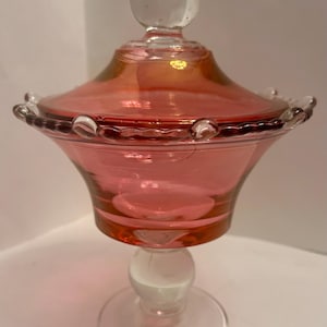 May include: A pink glass candy dish with a clear glass base and lid. The dish has a round base, a bulbous stem, and a flared bowl. The lid is domed with a clear knob. The rim of the dish is scalloped.