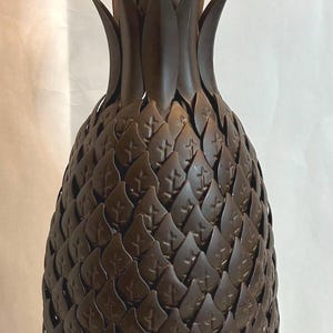 May include: A dark brown metal pineapple sculpture featuring a textured exterior composed of overlapping leaf-shaped scales. The top of the object displays a cluster of pointed, upright metal leaves, creating a realistic representation of a pineapple.