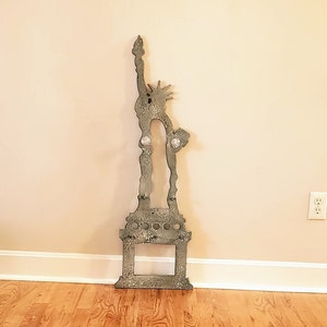 May include: A silver metal cutout of the Statue of Liberty standing on a wooden floor in front of a beige wall. A gray shag rug is in the foreground.