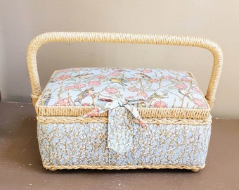 May include: A wicker sewing box with a floral patterned lid and a light blue and white floral design on the base. The box has a handle and is closed.