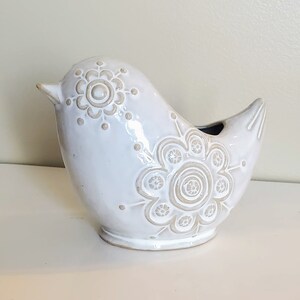 May include: White ceramic bird figurine with a decorative floral design. The bird has a rounded body and a long beak.