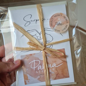 May include: A package of stationery wrapped in clear cellophane and tied with raffia. The package includes cards with the word "Surprise" and a hand-drawn illustration. A sticker with the words "Merci" and "Palma Atelier" is also visible.