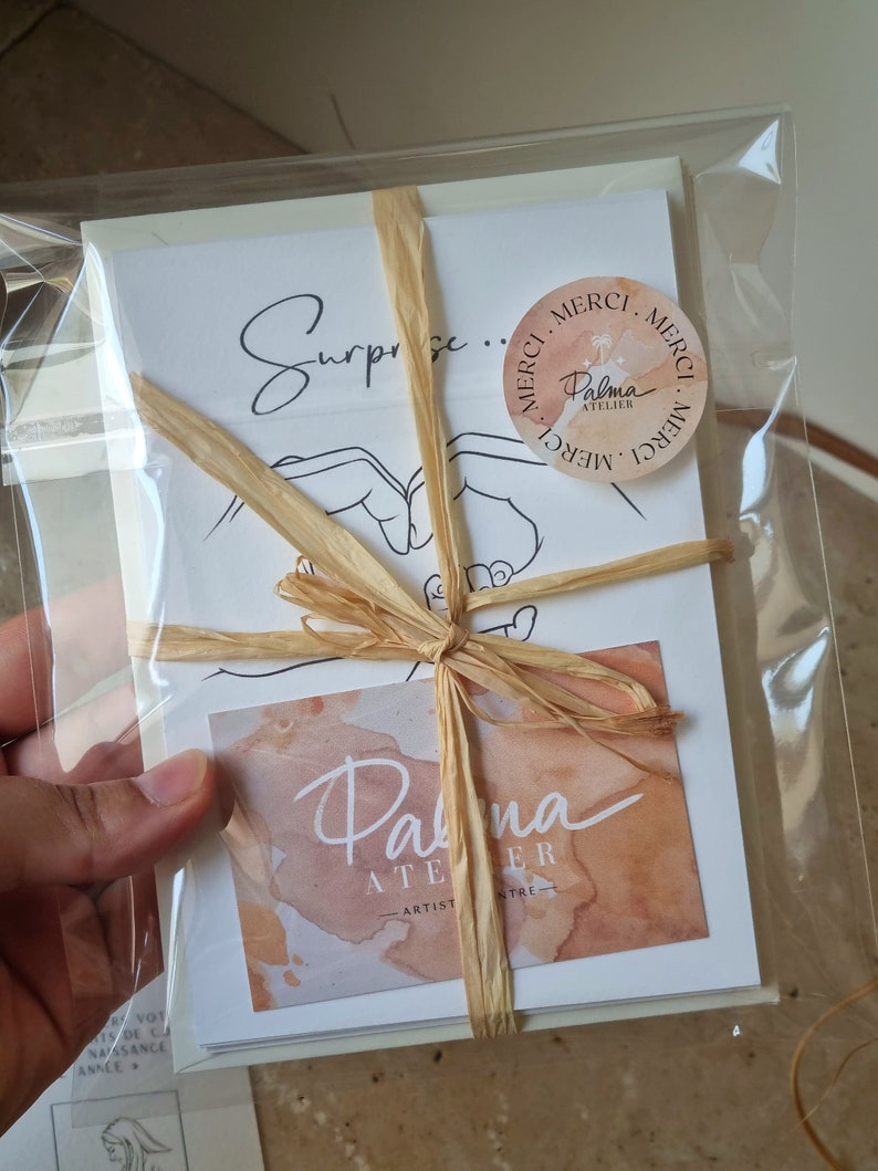 May include: A package of stationery wrapped in clear cellophane and tied with raffia. The package includes cards with the word "Surprise" and a hand-drawn illustration. A sticker with the words "Merci" and "Palma Atelier" is also visible.