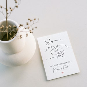 May include: A white card with the text "Surprise..." and a line drawing of hands forming a heart around a baby's feet. The card also includes the text "MINI NOUS ARRIVE BIENT&Ocirc;T Marine & Pedro" and a small pink heart.