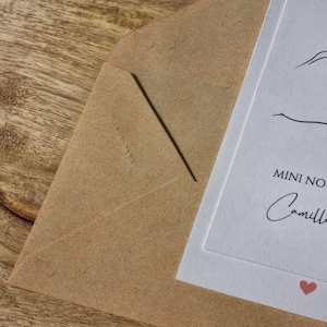 May include: A close-up of a kraft paper envelope with a white card inside. The card has the words "MINI NOTE" and the name "Camille" in script, with a small red heart. The envelope is on a wooden surface.