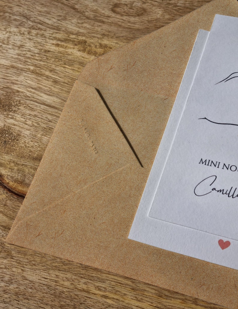 May include: A close-up of a kraft paper envelope with a white card inside. The card has the words "MINI NOTE" and the name "Camille" in script, with a small red heart. The envelope is on a wooden surface.