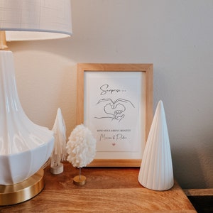 May include: A framed print with the text "Surprise..." and "Mini nous arrive bient&ocirc;t" in black script, with a heart illustration. A white lamp, decorative trees, and a pom-pom ornament sit on a wooden surface.