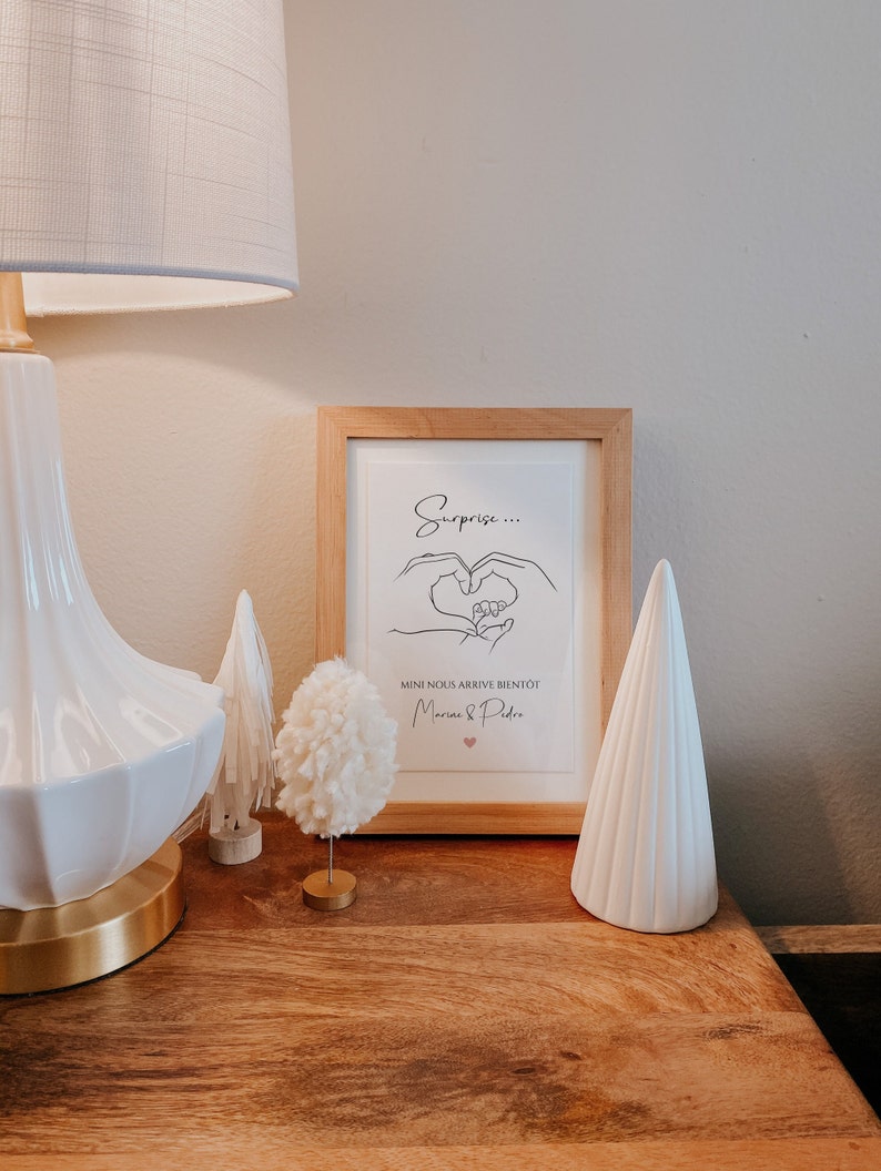 May include: A framed print with the text "Surprise..." and "Mini nous arrive bient&ocirc;t" in black script, with a heart illustration. A white lamp, decorative trees, and a pom-pom ornament sit on a wooden surface.