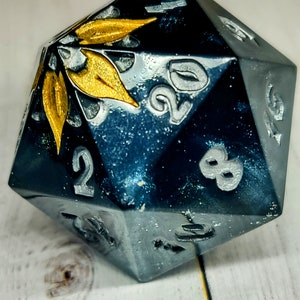 May include: A black and gold 20-sided die with a leaf design. The die is made of a sparkly material and has silver numbers.