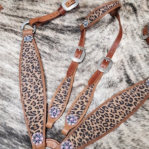 May include: A set of brown leather horse tack with a leopard print design. The tack includes a headstall, breast collar, and reins, all adorned with silver buckles and decorative jewels. The background is a mottled brown and gray pattern.