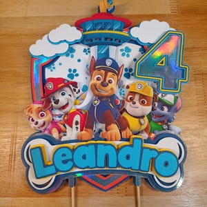Puede incluir: Adorno de pastel colorido con los personajes de la Patrulla Canina, el número 4 y el nombre "Leandro". El diseño incluye a Chase, Marshall, Rubble, Skye y Rocky. El adorno está montado en palitos de madera.