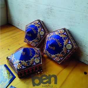 May include: Three blue and gold hexagonal boxes with the text "Chocolate Frog" are displayed on a wooden surface. The boxes have ornate designs and are accompanied by matching cards. The boxes are from "ben Papelería y Festejos".