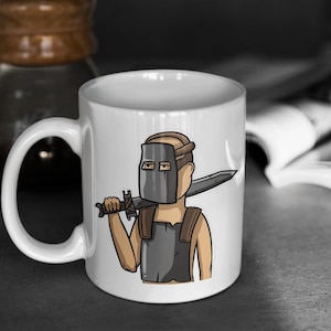 Puede incluir: Taza de cerámica blanca con una ilustración de dibujos animados de una persona con una máscara de metal y sosteniendo una espada. La persona lleva una placa de pecho de metal y tiene el pelo castaño.