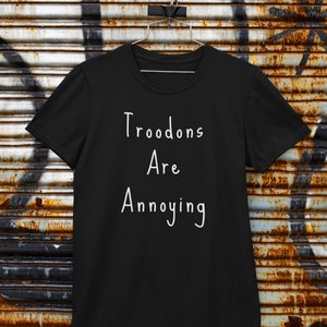 Może przedstawiać: Czarny t-shirt z białym napisem "Troodons Are Annoying" wiszący na rdzewiejącym metalowym tle.