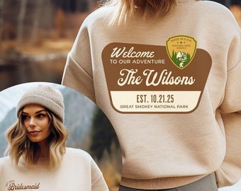 Op de afbeelding: Beige sweatshirt met een aangepast ontwerp op de rug. Het ontwerp bevat de tekst "Welcome to our Adventure", "The Wilsons" en "Est. 10.21.25". Een kleinere afbeelding van een berg en bomen is ook inbegrepen. Het sweatshirt wordt getoond voor een berg en een meer.