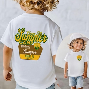 May include: A white t-shirt with a graphic that says "Camp Titi Summer Camp Camper Miami, FL Est. 2020" in green, yellow, and brown. The shirt is worn by a young child wearing a white baseball cap and denim shorts.