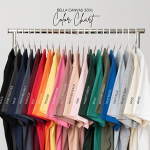 Puede incluir: Una carta de colores de camisetas Bella Canvas 3001 en varios colores, incluyendo negro, azul marino, rojo, amarillo, rosa, verde y azul. Cada camiseta est&aacute; etiquetada con su nombre de color, mostrada en un estante de metal.