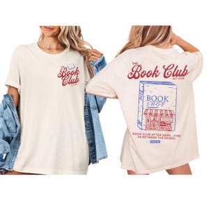 K&ouml;nnte beinhalten: Cremefarbenes T-Shirt mit "The Book Club" in roter Schrift und einer kleinen Buchgrafik. Auf der R&uuml;ckseite befindet sich eine Buchladen-Illustration mit dem Text "BOOK SHOP" und "BOOK CLUB AFTER DARK...FIND US BETWEEN THE PAGES!"