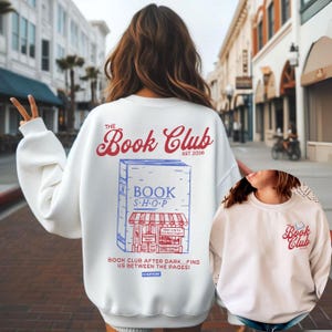K&ouml;nnte beinhalten: Wei&szlig;es Sweatshirt mit dem Schriftzug "The Book Club" in roter Schrift und einer blau-roten Buchladen-Grafik. Auf der R&uuml;ckseite des Sweatshirts steht der Text "Book Club After Dark... Find Us Between The Pages!" und "Everyday".