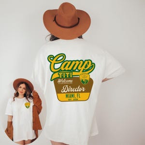 Puede incluir: Camiseta blanca con un dise&ntilde;o de Camp Titi Director en verde, amarillo y marr&oacute;n. El dise&ntilde;o incluye el texto "Welcome to our adventure" y "Miami, FL Est. 2026". Una versi&oacute;n m&aacute;s peque&ntilde;a del dise&ntilde;o est&aacute; en la parte delantera.